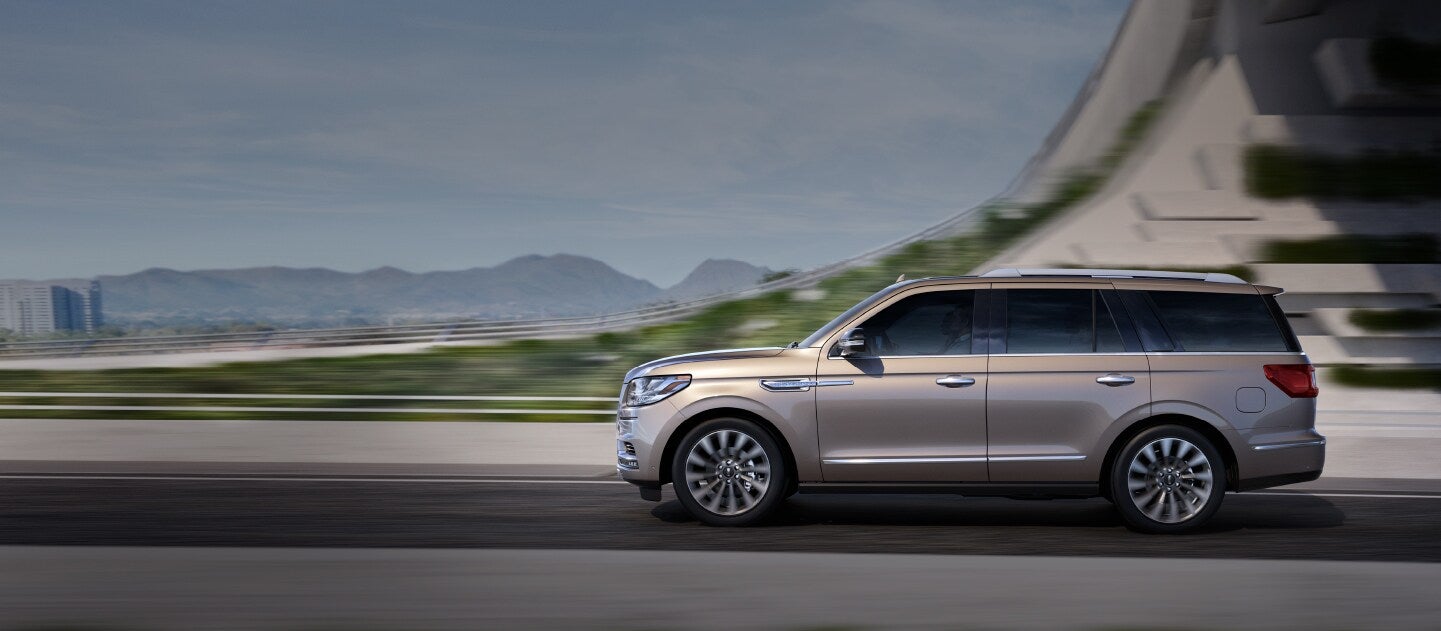 A Lincoln Navigator in Iced Mocha is emerging from the shadow of an overpass with its exterior curves lit by the light of a bright day.