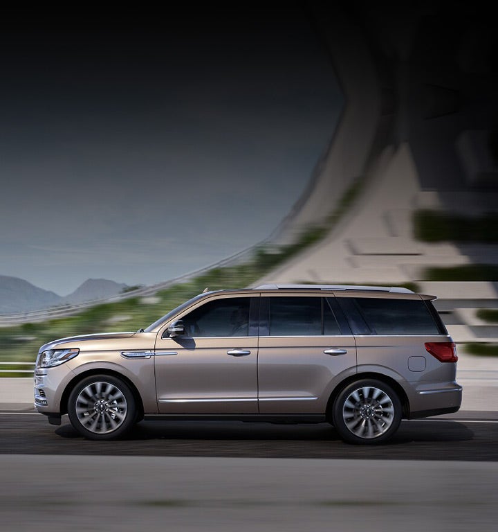 A Lincoln Navigator in Iced Mocha is emerging from the shadow of an overpass with its exterior curves lit by the light of a bright day.
